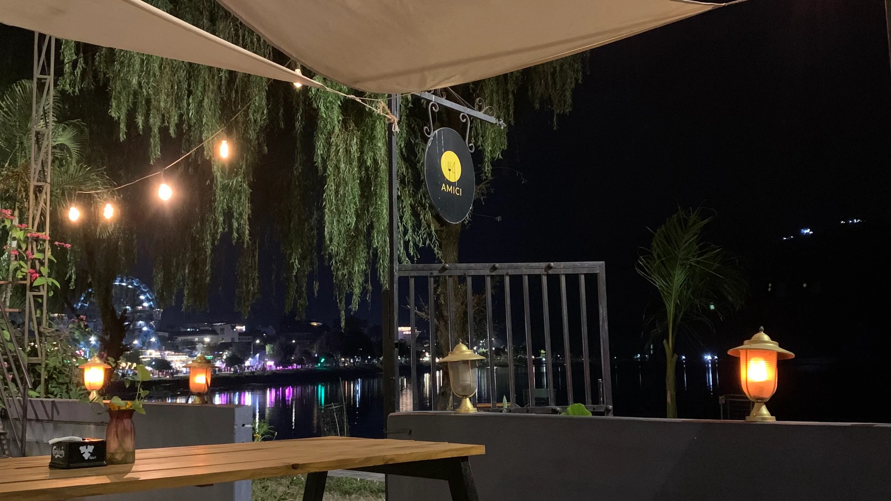 Amici exterior at night with lit AMICI sign, string lights, lanterns and lake reflections