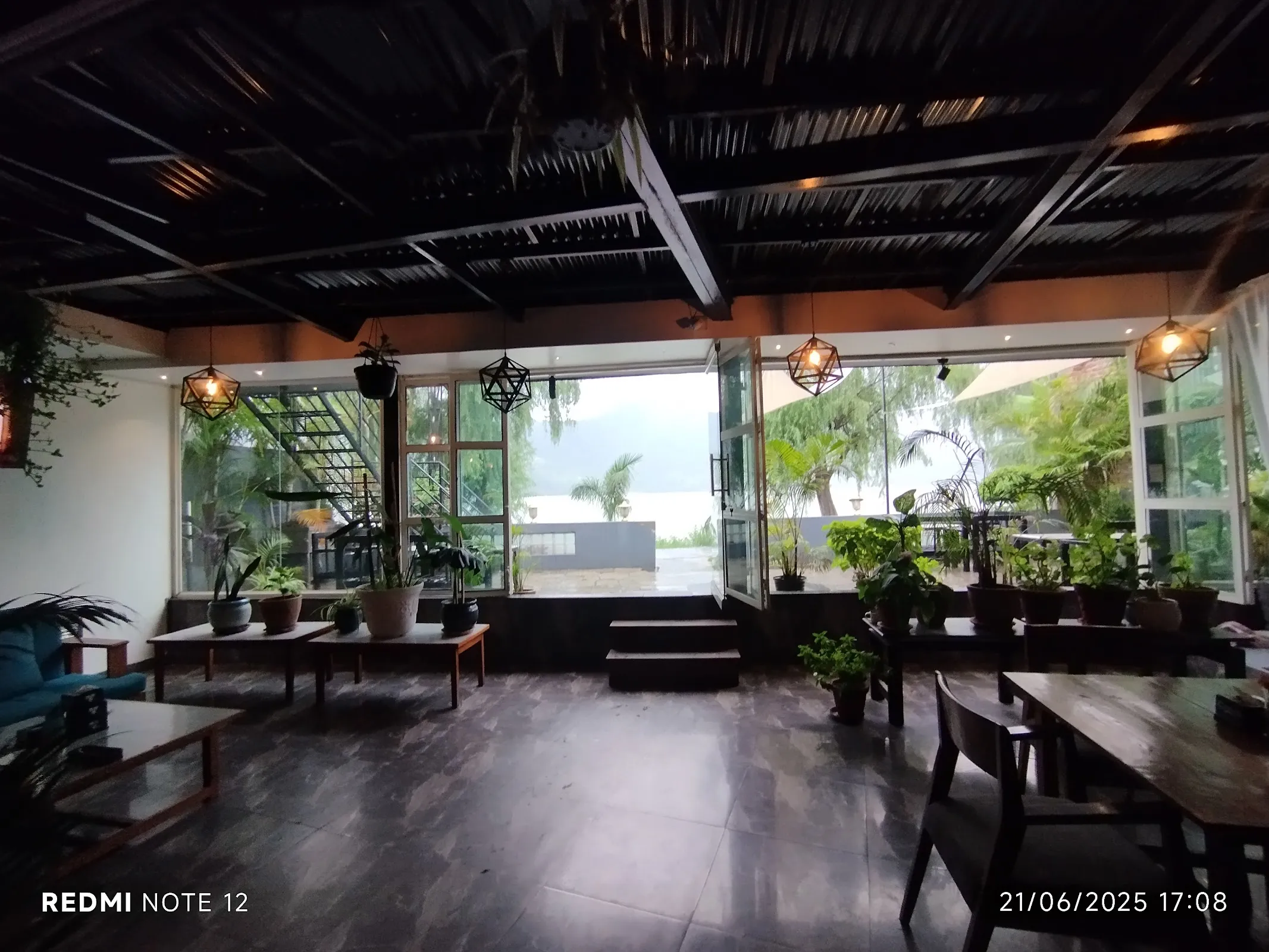 Wide interior view of Amici with open ceiling, exposed wooden beams, hanging plants and pendant lights — opens onto the lake