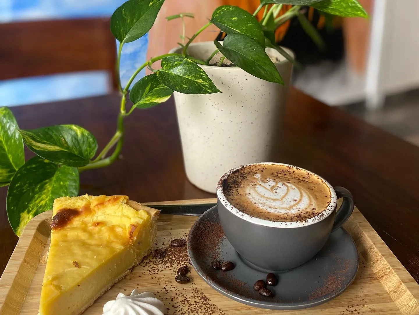 A slice of lemon tart on a wooden tray beside a cappuccino with foam art and a green plant
